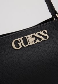 Sac à main texturé noir avec un logo Guess en métal doré et une poignée en cuir attachée par un anneau métallique.