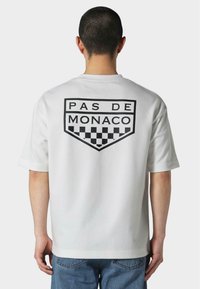 Bílý krátký rukáv tričko s černou grafikou "PAS DE MONACO" a kostkovaným vzorem na zadní straně. Měkký bavlněný materiál.