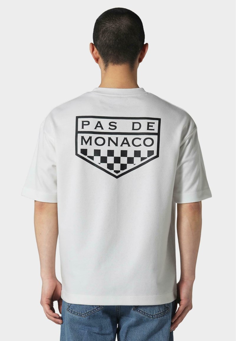 Bílý krátký rukáv tričko s černou grafikou "PAS DE MONACO" a kostkovaným vzorem na zadní straně. Měkký bavlněný materiál.