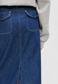 Saia de ganga em azul-escuro, com duas bolsas traseiras com fechos de botão, costura branca em contraste e uma fenda central nas costas.