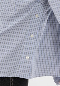 Ανοιχτό μπλε καρό πουκάμισο με άσπρα κουμπιά, φορεμένο πάνω από μαύρα τζιν παντελόνια, δείχνοντας λεπτομέρειες από τα κουμπιά και την υφή του υφάσματος.