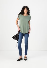 Green short-sleeve top, slim-fit blue jeans, black patterned handbag with ruffled details, black flats with round accents, white background.