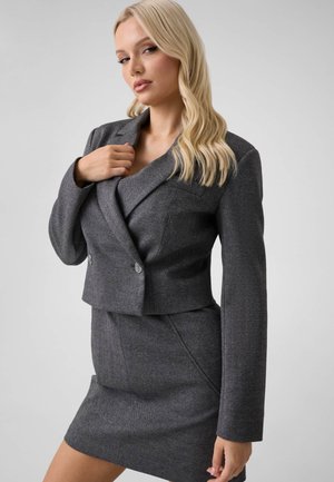 Mujer rubia con un blazer gris ajustado y falda a juego, posando con una mano ajustando la solapa del blazer sobre un fondo claro y liso.