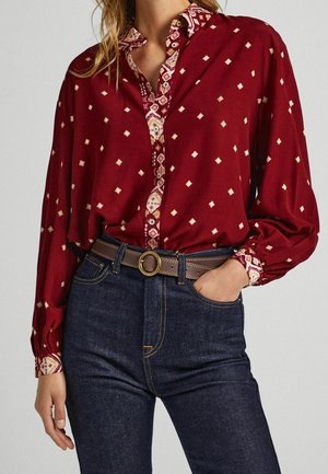 Donna che indossa una camicetta a maniche lunghe rossa con un motivo, infilata in jeans di denim scuro con una cintura marrone con fibbia rotonda in ottone.