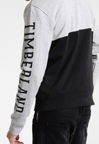 Grijs-zwart sweatshirt met een kleurblokontwerp, met "TIMBERLAND" in vetgedrukte letters langs de linkermouw. Zachte textuur en ronde hals.