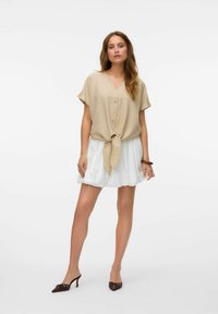 Beige blouse met korte mouwen en een v-hals, met een strik aan de voorkant, gecombineerd met een witte uitlopende rok. Bruine haksandalen maken de outfit compleet.
