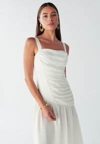 Bílé šaty bez rukávů s mačkaným živůtkem a splývavou sukní. Mají jemnou texturu a široká ramínka. Na zápěstí zlatý náramek.