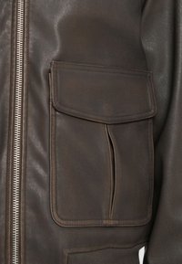 Lähikuva tummanruskeasta nahkatakista, jossa on suuri etutasku, jossa on läppä ja näkyvä pystysauma, sekä hopeinen vetoketju sivussa.