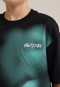 Juodas trumpomis rankovėmis t-shirts su gradientiniu teal taškų raštu ir baltu "SALTYDOG" logotipu priekyje. Medvilnė, klasikinis fasonas.