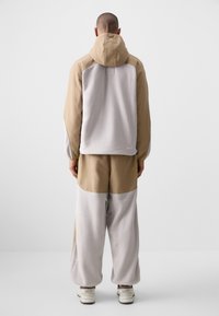 Khaki og grå to-farget hettejakke med en myk, teksturert overflate og avslappet passform, kombinert med matchende bukser som viser en løs design.