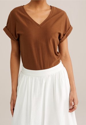 Bruine top met V-hals en textuur, met opgerolde mouwen, gecombineerd met een vloeiende witte rok. De stof heeft een subtiel patroon en een lichte val.