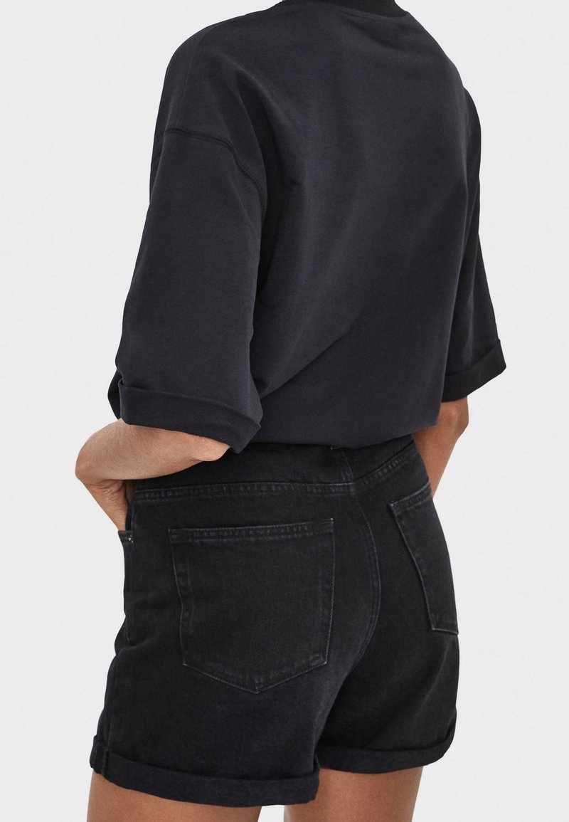 Personne portant une chemise noire aux manches retroussées et un short en jean noir avec des poches arrière, les mains reposant près des poches, fond clair uni.