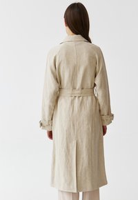 Beige linen trench coat with a belted waist, long sleeves, and buttoned cuffs. Features a vent at the back and a classic collar design.