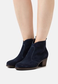 Botines de ante azul marino con forma cónica, paneles laterales curvados y un tacón bajo de madera. Presentan una textura suave y un diseño elegante.