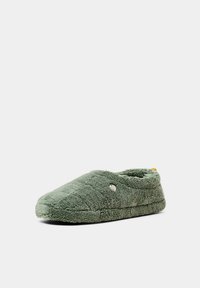 Chausson doux, vert, fermé à l’arrière, en tissu peluche avec une couture décorative texturée et un petit logo rond sur le côté.