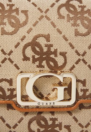 Tissu tissé beige et marron avec motif monogramme répété "G" et "S" et un emblème métallique doré "G" étiqueté "GUESS" sur un sac.