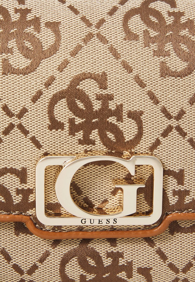 Tissu tissé beige et marron avec motif monogramme répété "G" et "S" et un emblème métallique doré "G" étiqueté "GUESS" sur un sac.