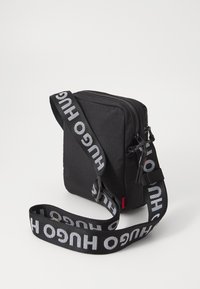 Borsa a tracolla nera in tessuto con chiusura a zip, forma piatta e spallaccio nero a contrasto con la scritta "HUGO" in grandi lettere bianche.