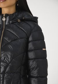 Giacca a vento nera con cappuccio, design trapuntato, minuteria dorata e chiusura con zip. Texture liscia e lucida con una forma sagomata.