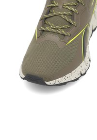 Zielono-oliwkowy but sportowy z teksturowaną cholewką z siateczki, żółtymi akcentami, białą podeszwą środkową z plamkami oraz czarną gumową podeszwą zewnętrzną.