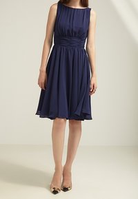 Granatowa sukienka bez rękawów z plisowanym fasonem, marszczoną talią i zwiewnym dołem. Połączona ze złotymi butami na obcasie. Gładka tekstura materiału.