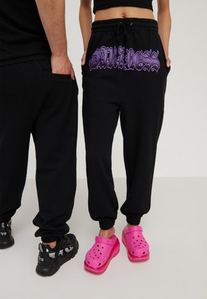 Zwei Personen tragen schwarze Hosen; eine hat vorne lila "DARKNESS"-Text, die andere trägt schlichte schwarze Hosen, leuchtend pinke Clogs sind sichtbar.