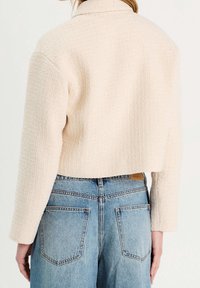 Veste courte texturée couleur crème avec col et manches longues, portée sur un jean denim bleu clair taille haute, dévoilant des détails dans le dos.