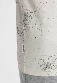 Camiseta beige clara con diseño de texto negro en diferentes tamaños, con una etiqueta que dice "ambre wear" en la costura lateral izquierda. Material suave, corte casual.