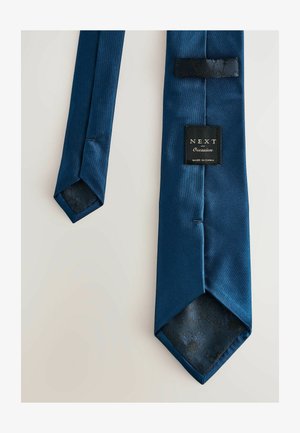 Tumši zils satīna kakla saite ar strukturētu melnas ziedu oderi, uz kuras ir uzraksts "NEXT Occasion" un cilpa, lai to nostiprinātu.