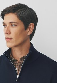Marineblaues Zip-Pullover mit geripptem Kragen über einem braunen Karohemd. Das Material scheint strukturiert zu sein, mit einem glatten Reißverschlussdetail am Hals.