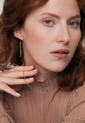 Złote kolczyki łańcuszkowe z niebieskimi akcentami, pasujący złoty naszyjnik łańcuszkowy oraz prosty złoty pierścionek. Modelka ma na sobie teksturowaną brązową bluzkę.