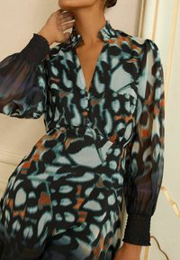 Bluză evazată din material transparent, cu un model abstract în culorile negru, albastru și portocaliu. Prezintă un guler volant și manșete cu elastic. Nasturi vizibili.