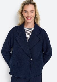 Námornícky modrý štruktúrovaný blazer s strapcovým lemom, klopovým golierom, uzatváraním na jeden gombík a dvomi prednými vreckami. Pod ním je košeľa s vertikálnym prúžkom.