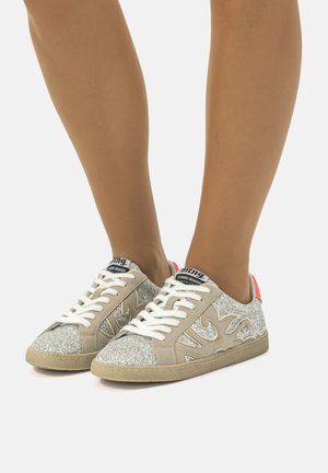 Zapatillas de deporte beige brillantes con cordones blancos, detalles grises y lengüeta en rosa. Presentan una parte superior texturizada y un patrón distintivo en los laterales.