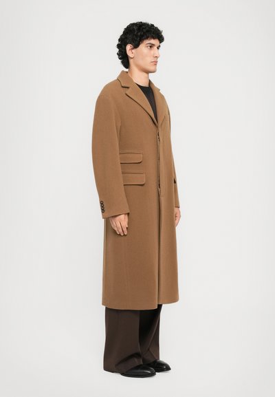 Man standing sideways wearing a long brown coat over dark wide-leg pants and black shoes against a plain white background.