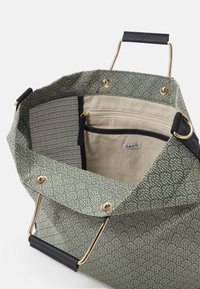 Zielona torba typu tote o teksturowanej powierzchni z złotymi akcentami, otwarta u góry i z czarnymi detalami. Zawiera beżową wewnętrzną kieszeń na zamek oraz metkę z logo.