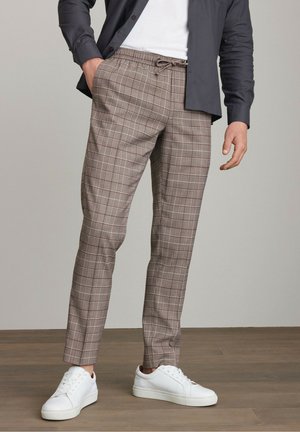 Hnědé kostkované kalhoty s elastickým pasem a stahovací šňůrkou, stylizované s bílými teniskami. Materiál má lehkou strukturu.