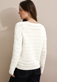 Pull en laine blanc avec un motif de rayures texturé et une encolure large. Manches longues et coupe décontractée. Pantalon en denim foncé visible à la taille.