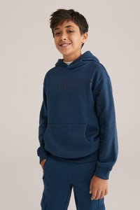 Temno modra kapuca s kenguru žepom, teksturirana tkanina in natisnjenim besedilom. V kombinaciji z enakimi temno modrimi hlačami, ki imajo cargo žepke.