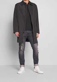 Mand iført en sort frakke over en sort skjorte, falmede slidte grå jeans rullet op ved anklerne og hvide sneakers, står op ad en ensfarvet væg.