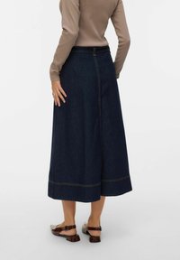 Tamsiai mėlynas džinsinis midi sijonas su tiesia siluetu, matomomis siūlių detalėmis ir išsišakojusia apačia. Derinamas su šviesios spalvos batais.