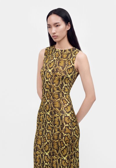 Femme aux longs cheveux noirs portant une robe sans manches, cintrée, avec un imprimé serpent jaune et marron sur un fond clair uni.