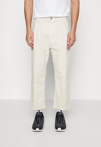 Beige beskurna byxor med knappstängning, framfickor och raka ben, kombineras med svarta sportskor med en texturerad design.