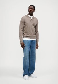 Καφετί πουλόβερ πόλο με μακριά μανίκια πάνω από μια λευκή μπλούζα, συνδυασμένο με φαρδιά μπλε τζιν και λευκά sneakers. Απλός σχεδιασμός και άνετη εφαρμογή.