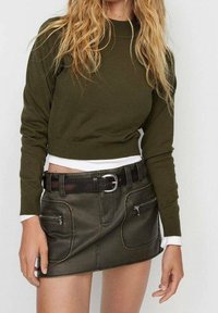 Olivgrüner, kurzer Pullover mit langen Ärmeln, über einem weißen Hemd getragen, kombiniert mit einem dunkelgrünen, ledernen Minirock mit zwei Reißverschlusstaschen.