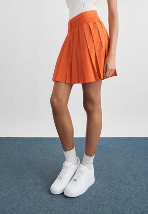 Femme portant une jupe plissée orange, des chaussettes blanches et des baskets blanches, debout sur un sol recouvert d'un tapis bleu, devant un mur blanc.
