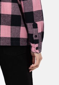 Camicia flanella a quadri rosa e nera con polsini bottonati e una texture liscia, accompagnata da un paio di pantaloni scuri e aderenti sullo sfondo.
