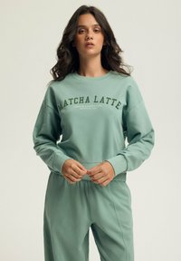 Miętowa bluza z napisem „MATCHA LATTE”, charakteryzująca się luźnym krojem, okrągłym dekoltem i długimi rękawami. Miękka w dotyku, zestawiona z pasującymi spodniami.