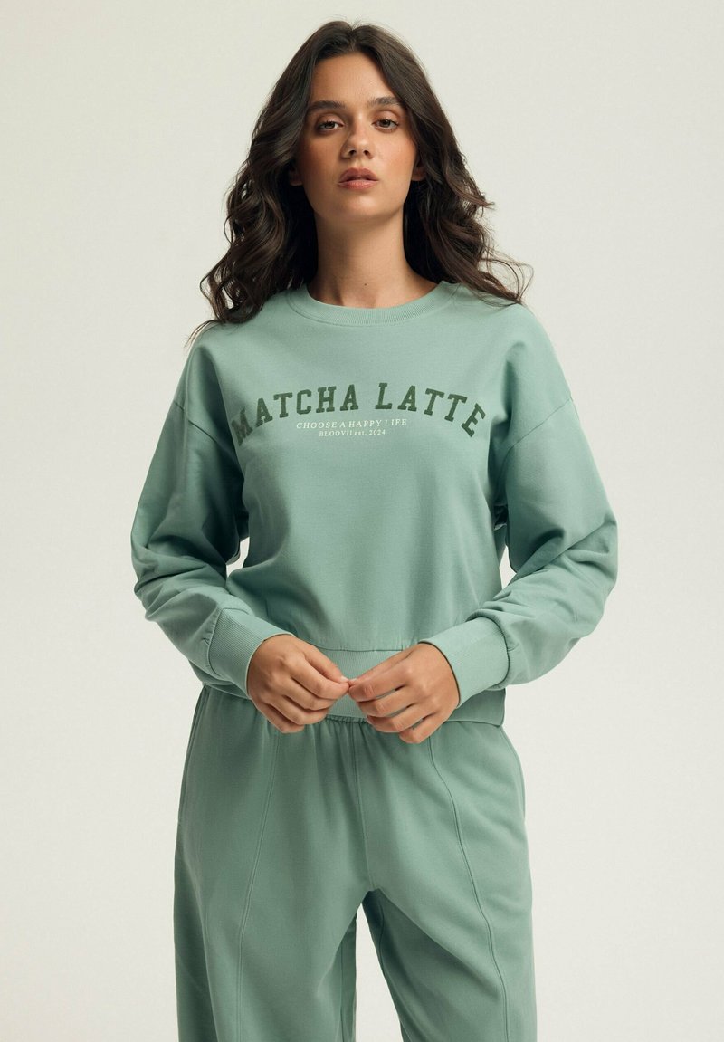 Miętowa bluza z napisem „MATCHA LATTE”, charakteryzująca się luźnym krojem, okrągłym dekoltem i długimi rękawami. Miękka w dotyku, zestawiona z pasującymi spodniami.