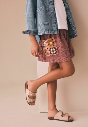 Copil purtând o jachetă din denim, cămașă albă, fustă roz texturată cu aplicatie croșetată și sandale bej cu cataramă, stând cu un picior ridicat.